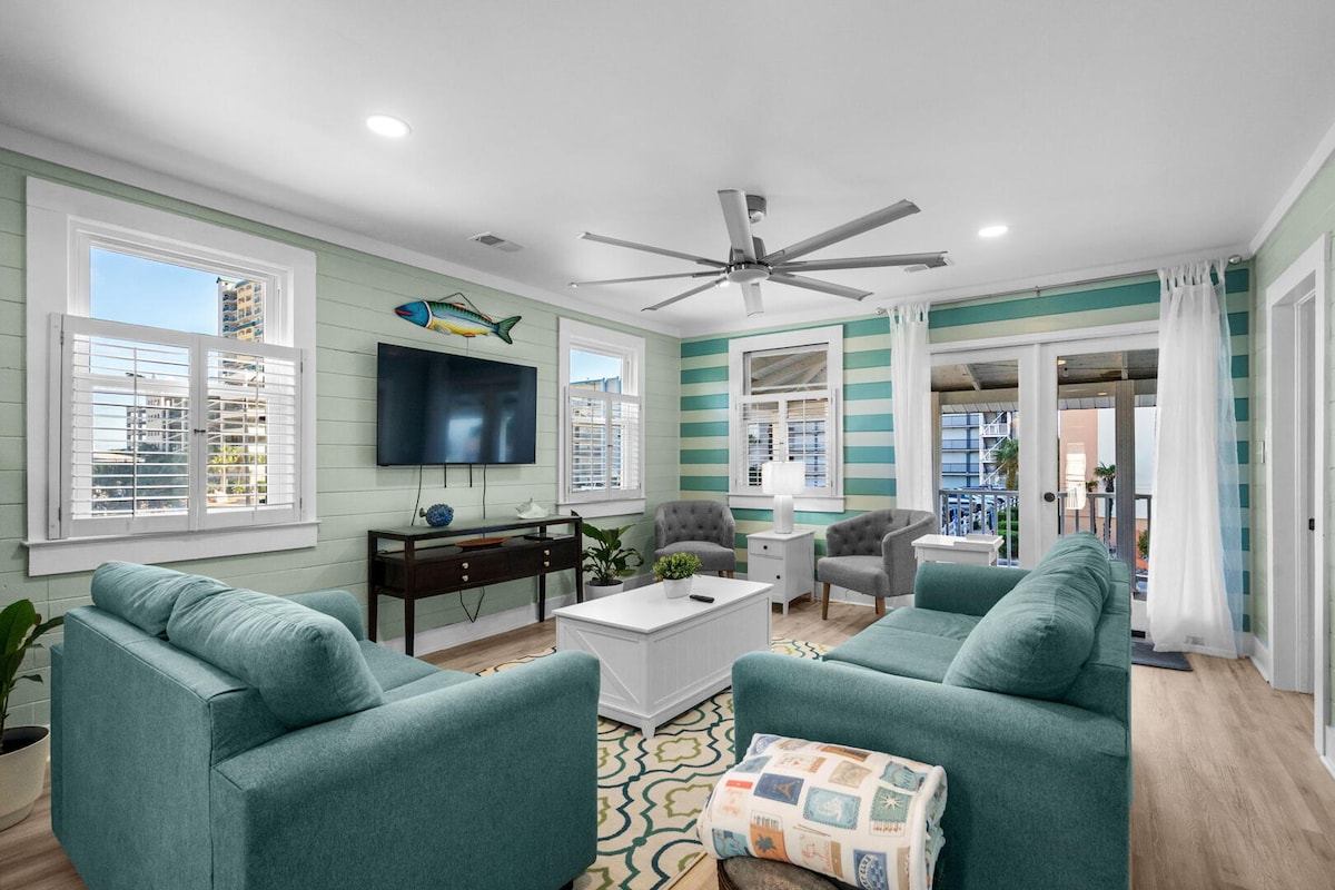 The open coastal living room with teal sofas, striped accent wall and French doors to the porch